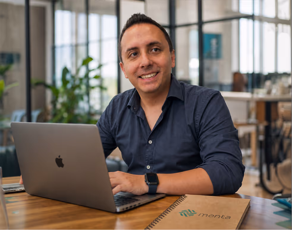 Luis Alfredo Ponce smiling in dark shirt working on a laptop at a wooden table with a notebook labeled Menta.